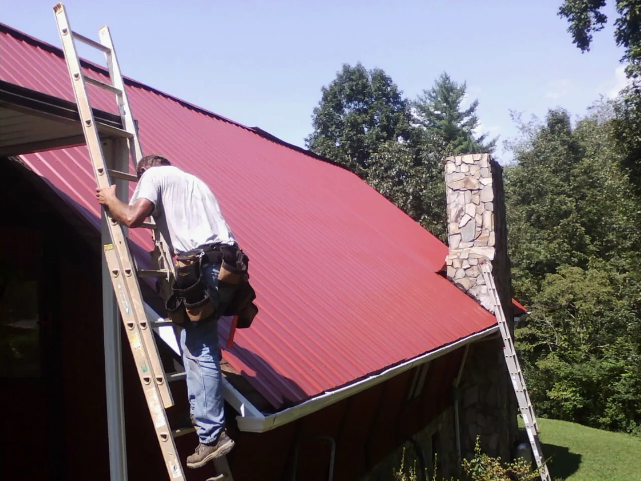Roofer on a ladder beside a red metal roof and stone chimney — Soffit & Fascia Repair by Roofing Brothers in North Bridgton