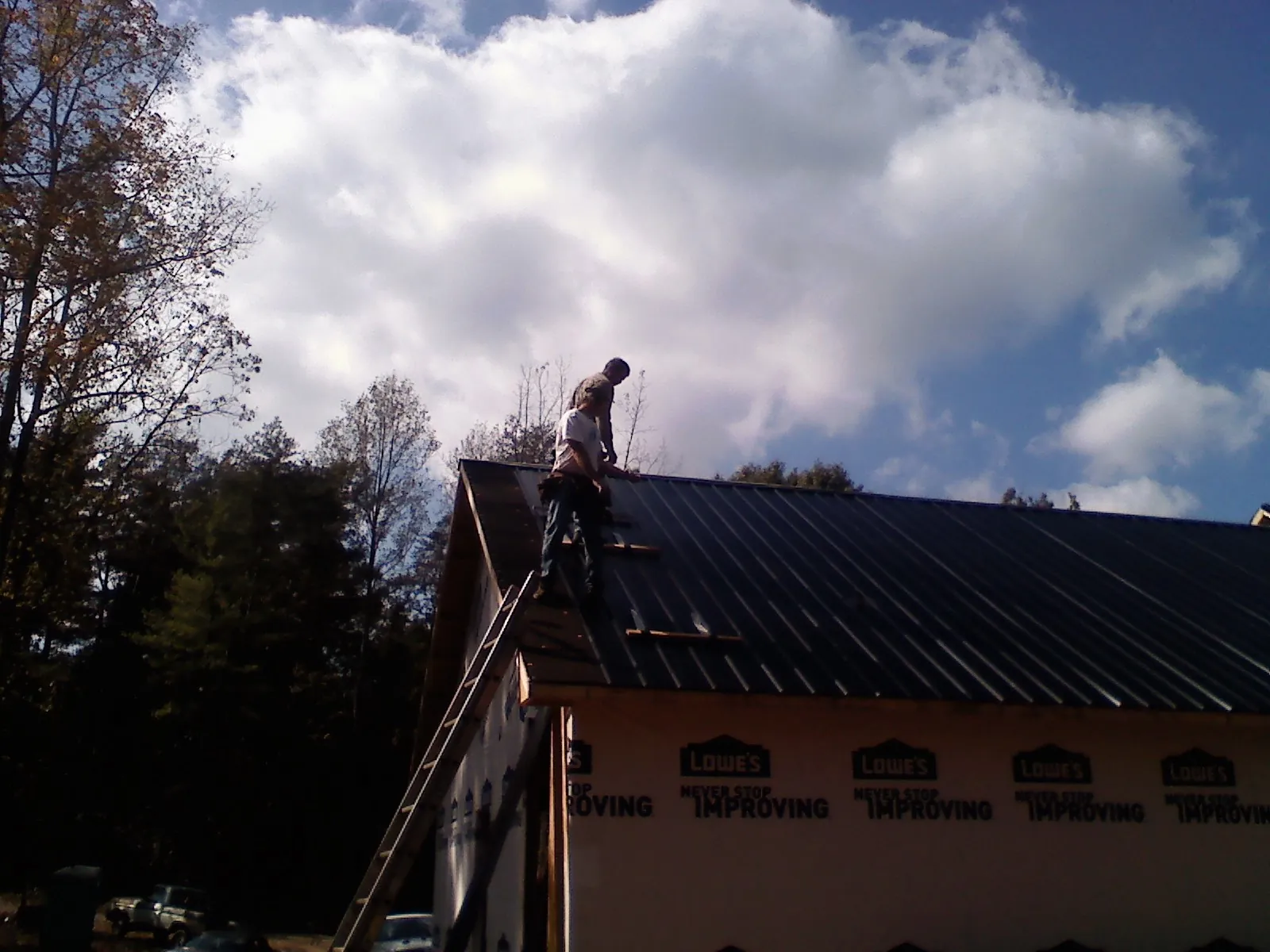 Roofer fastening dark metal panels high against a cloudy sky, from a Roof Inspection job in North Bridgton